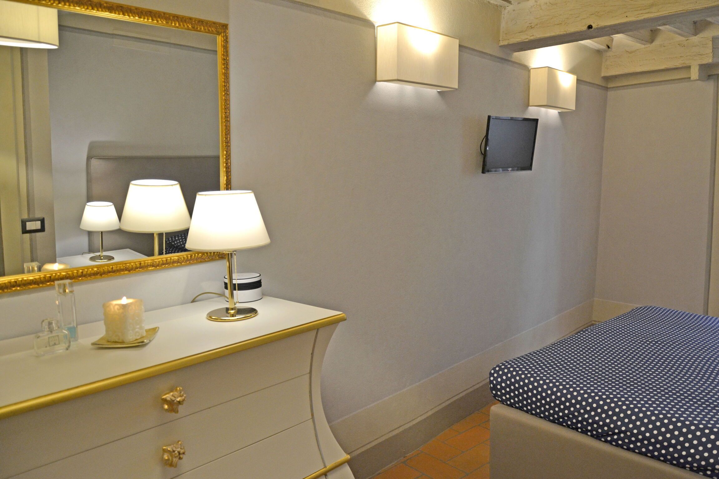 Quarto com cama, mesa de cabeceira com luminária, espelho e televisão na parede.