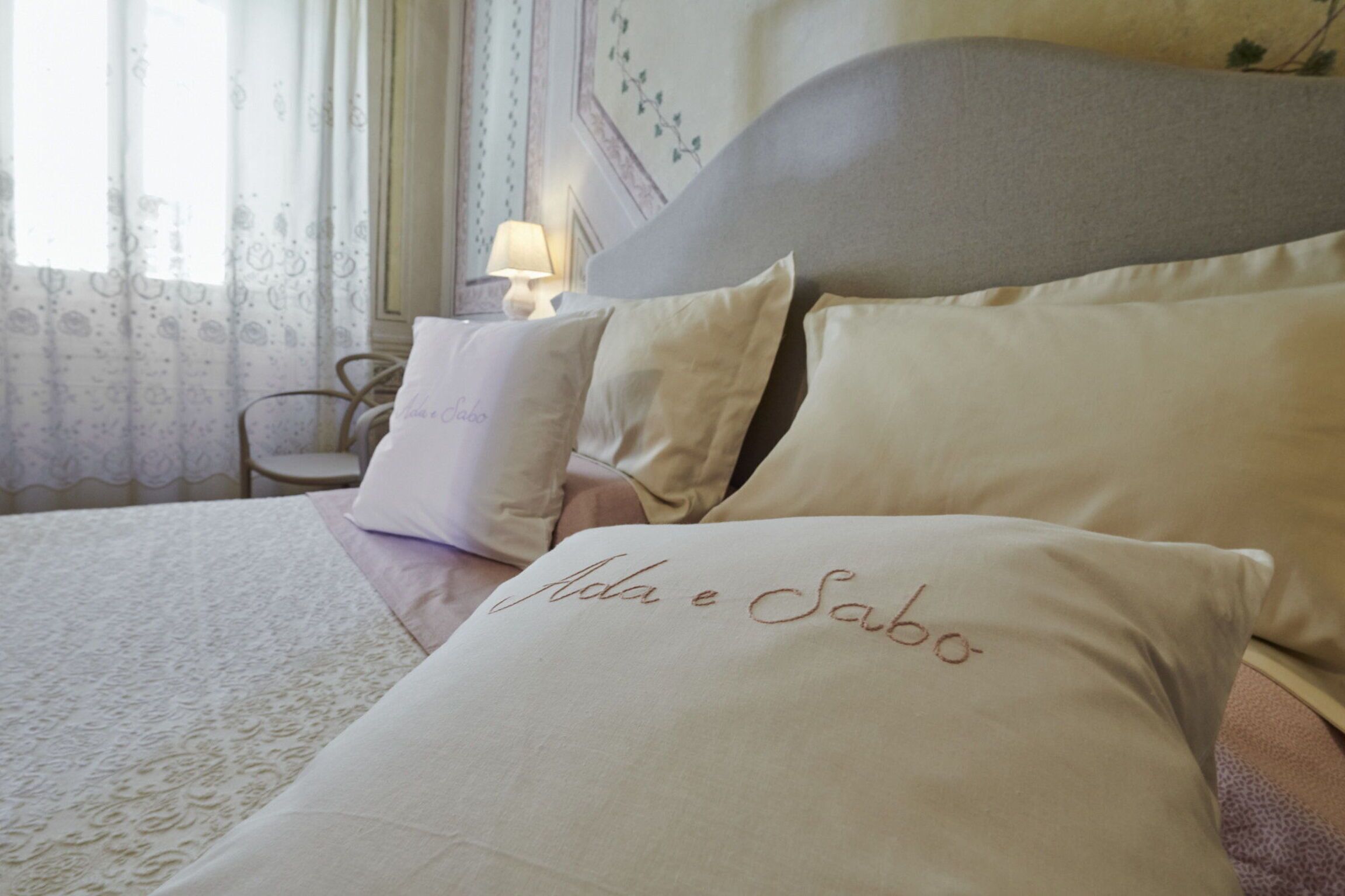 Una cama con almohadas que dicen 'Ada e Sabo', una lámpara en una mesita de noche y una silla en una habitación con cortinas estampadas.