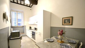 A compact kitchen with a dining area, a small refrigerator, a window, and a framed picture on the wall. - Appartamento La Piazzetta (Cortona)