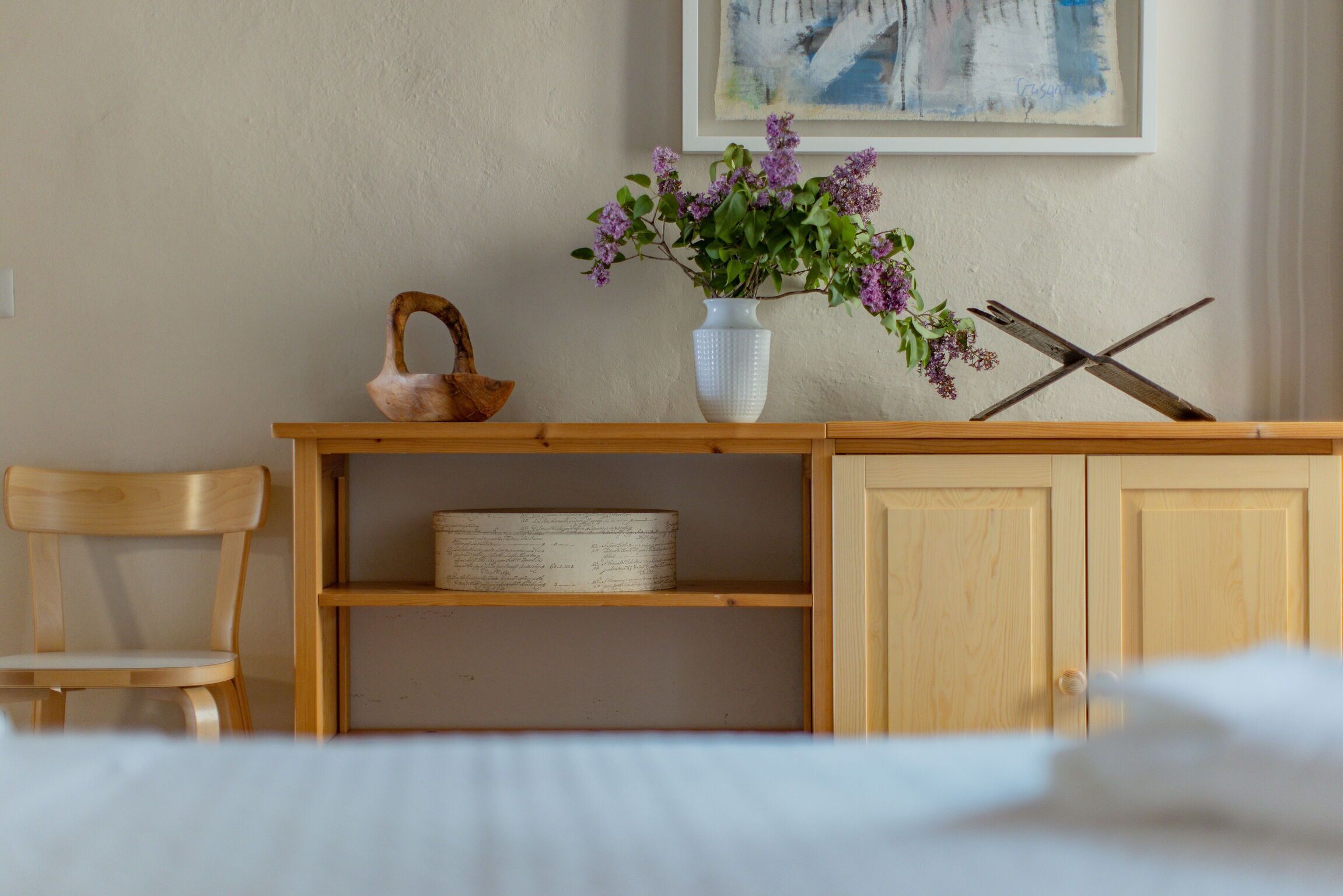 A wooden chair, a white vase with purple flowers, a basket, and a shelf with a box.