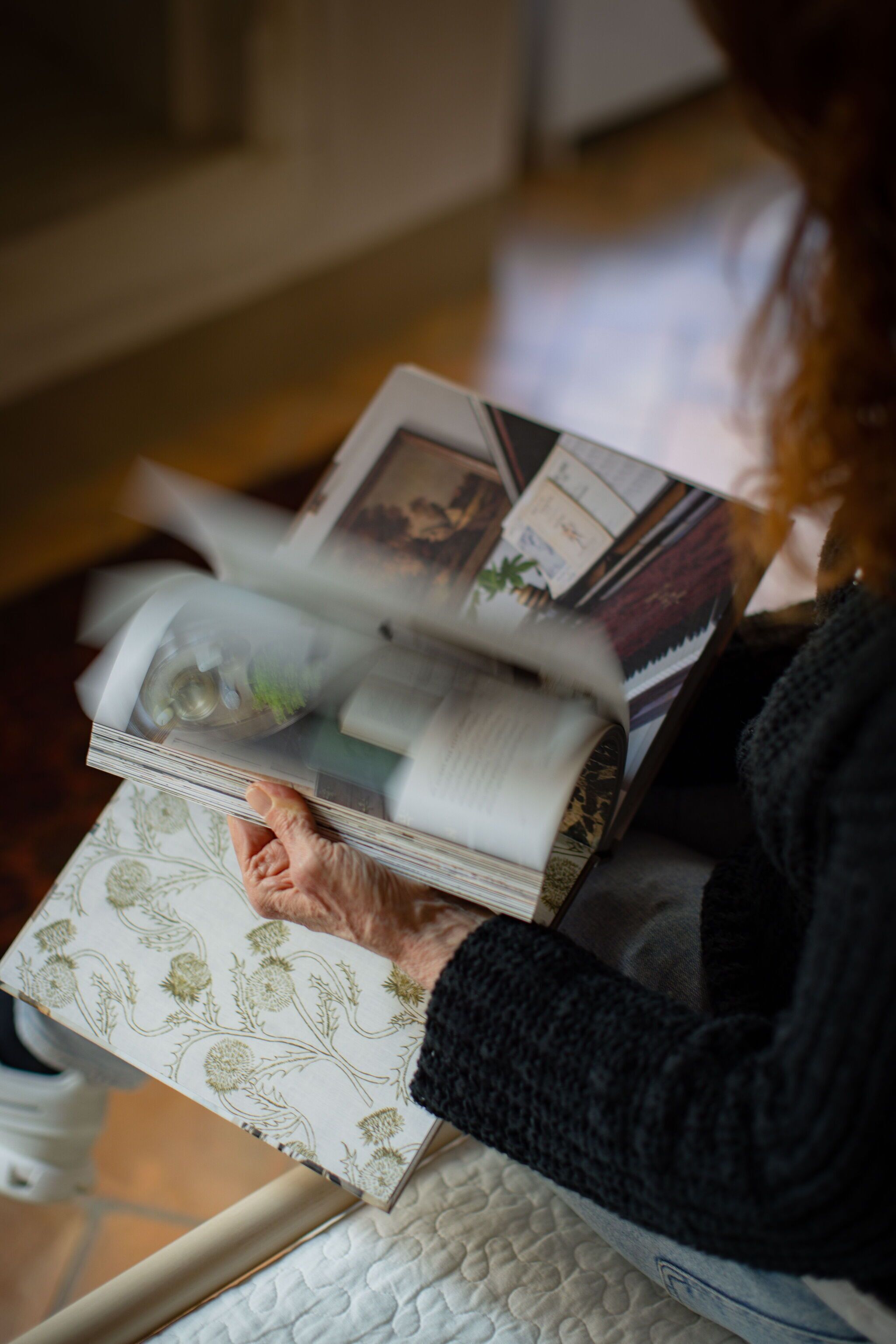 A person holding a magazine with a floral pattern, a quilted pillow, and a glass object on a table.