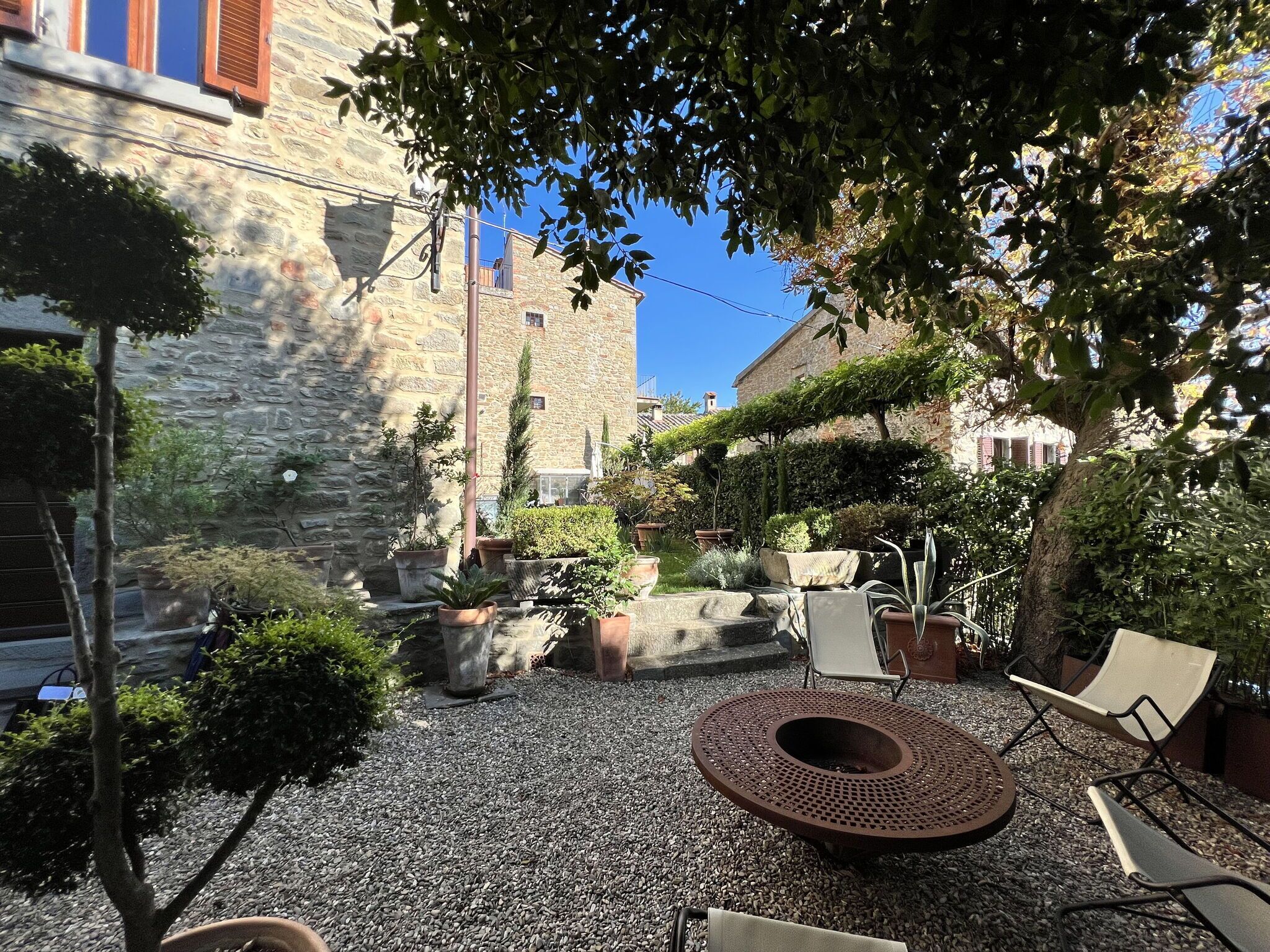 A courtyard with stone walls, potted plants, a circular fire pit, and outdoor seating.