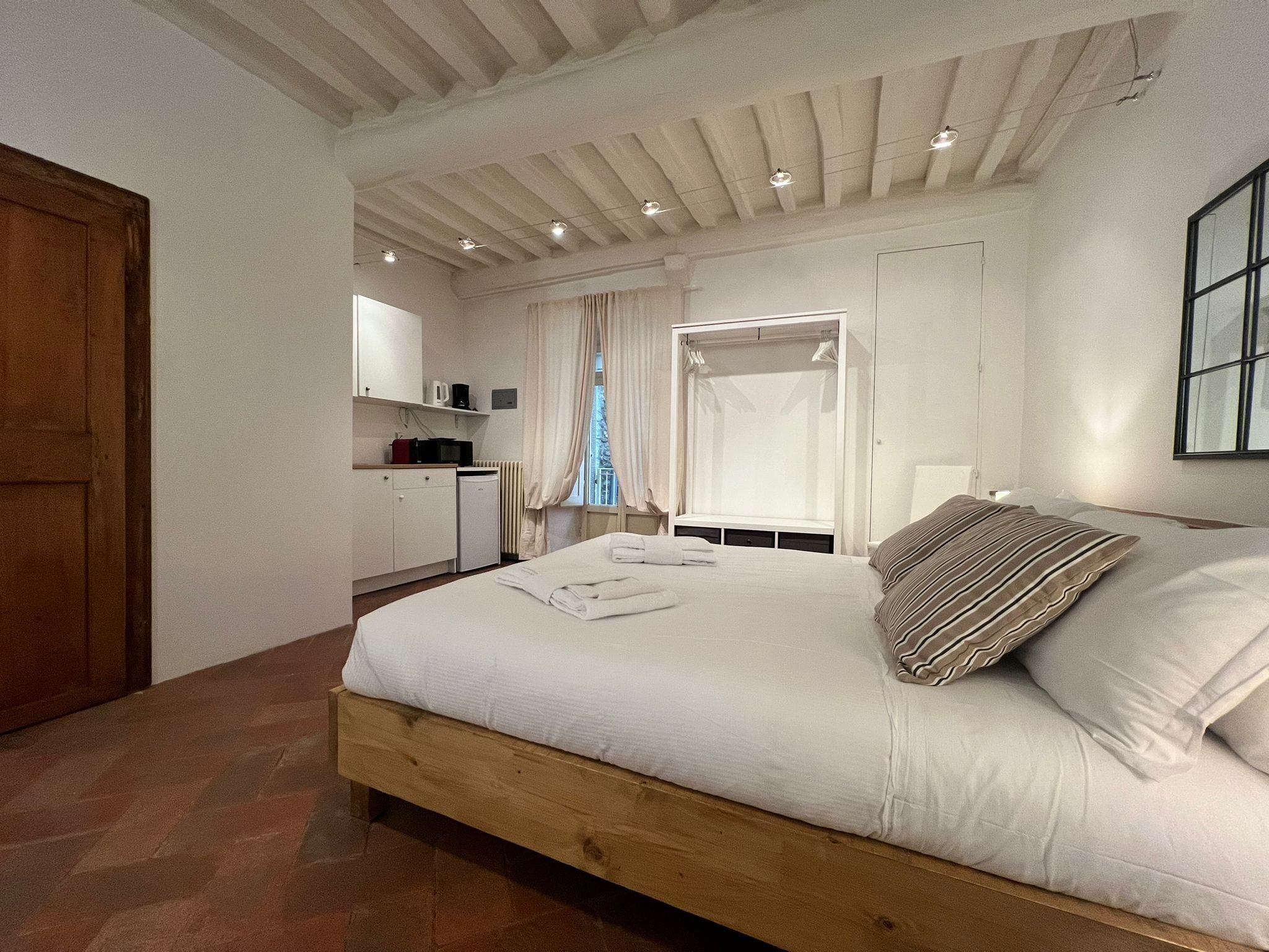 A bedroom with a wooden bed, a small kitchenette, and a large window with curtains.