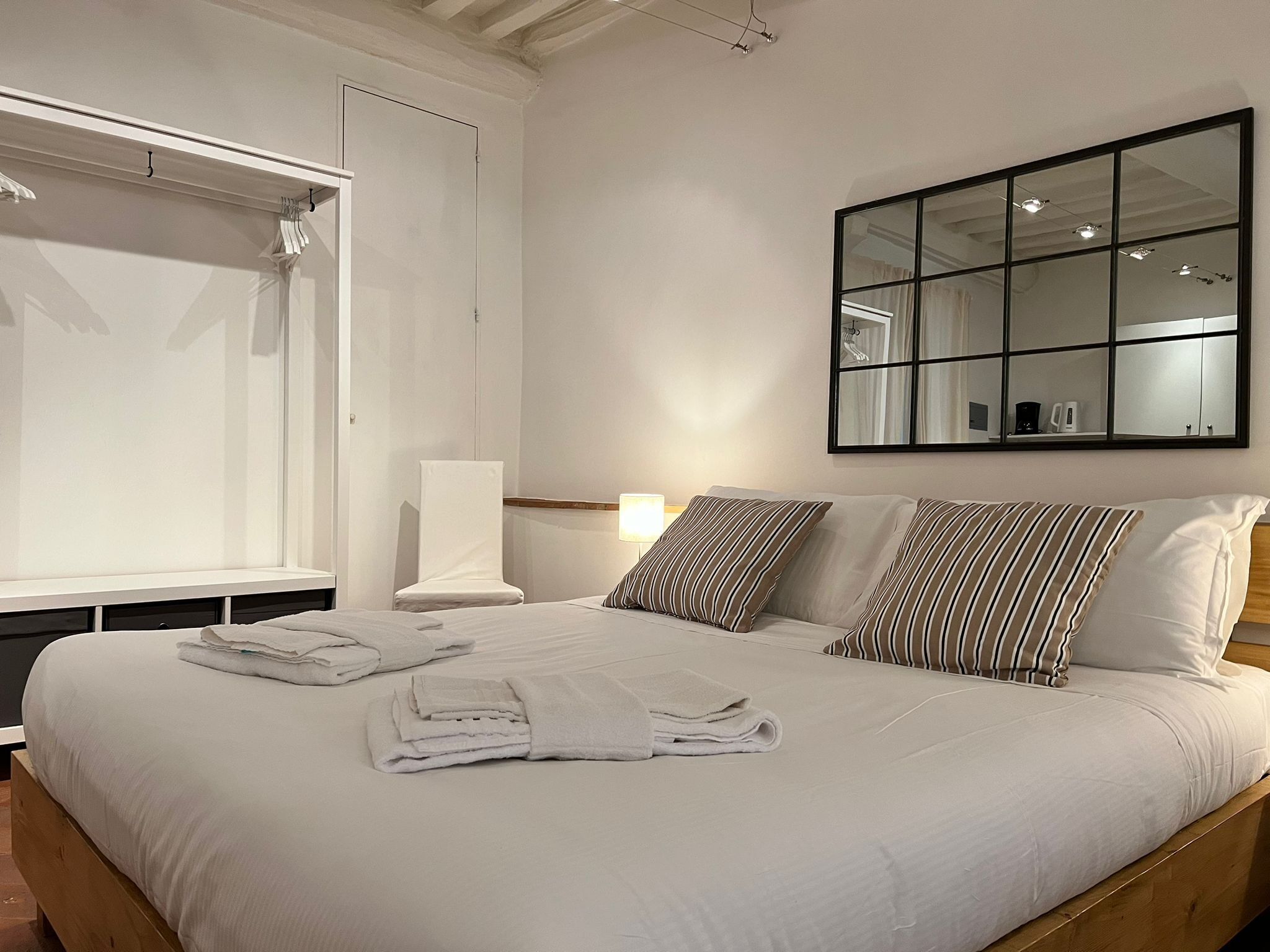 A neatly made bed with white linens and striped pillows, a bedside lamp, and a large mirror on the wall.