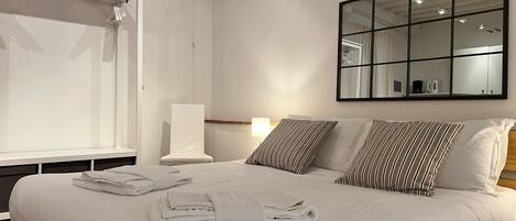 A neatly made bed with white linens and striped pillows, a bedside lamp, and a large mirror on the wall.