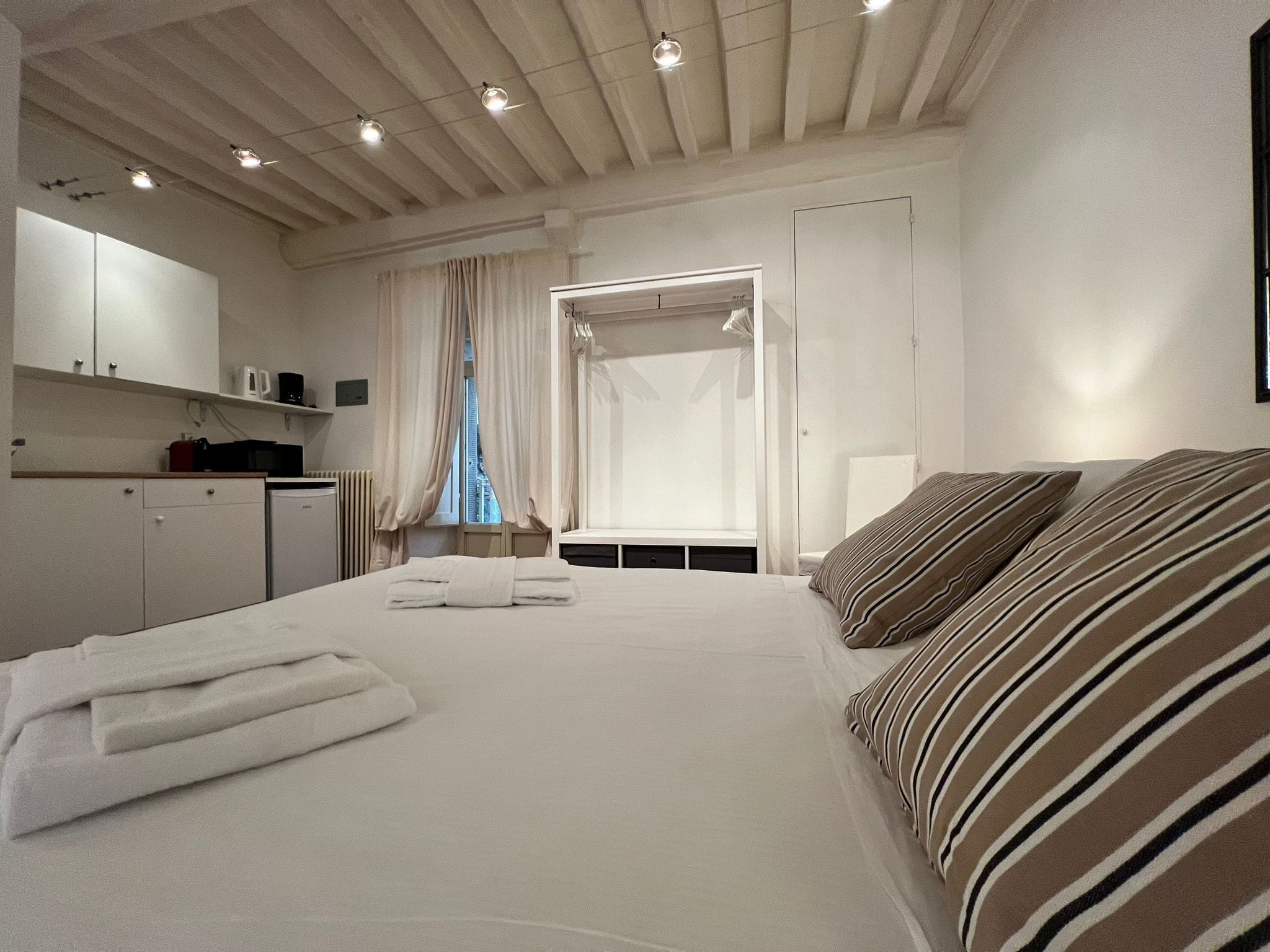 Un dormitorio con una cama grande, una mesita de noche, un armario, una ventana con cortinas y una zona de kitchenette.