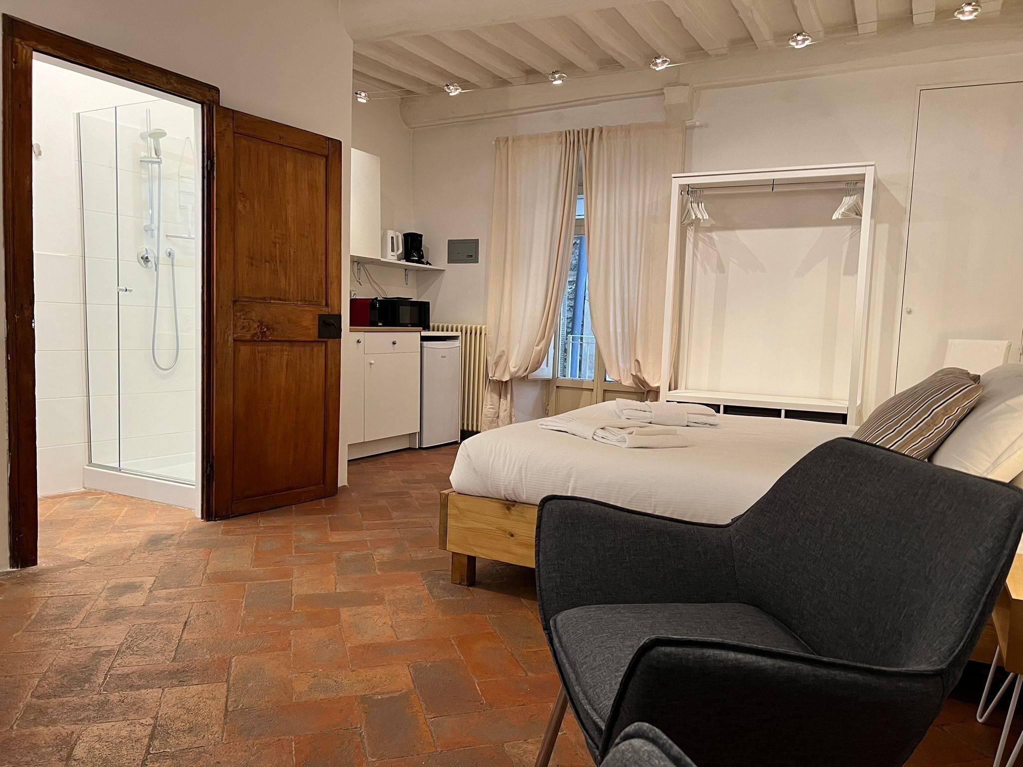 A bedroom with a bed, a chair, a wardrobe, a shower area, and a small kitchenette.