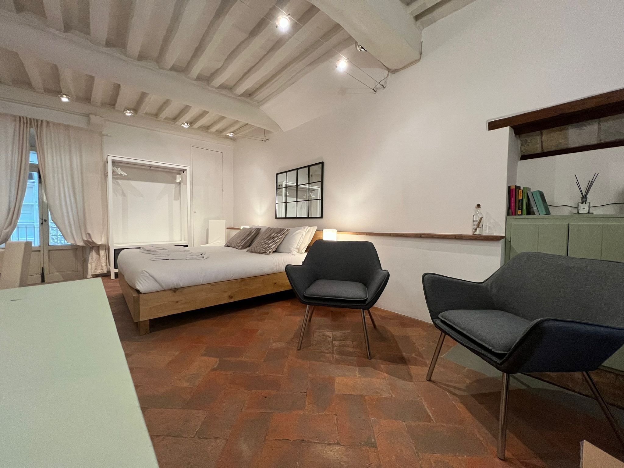 Una camera da letto moderna con un letto di legno, una scrivania, due sedie, uno specchio e un mobiletto verde pieno di libri.