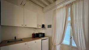 A compact kitchen with white cabinets, a built-in sink, a microwave, and a coffee maker. There is a window with white curtains and a radiator below. - Vicolo Della Scala Rooms - Il Sole (Cortona)