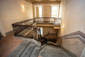 A grand staircase with ornate black railings, a stone floor, and a view of the upper floor through a window. - Appartamento Vagnucci (Cortona)