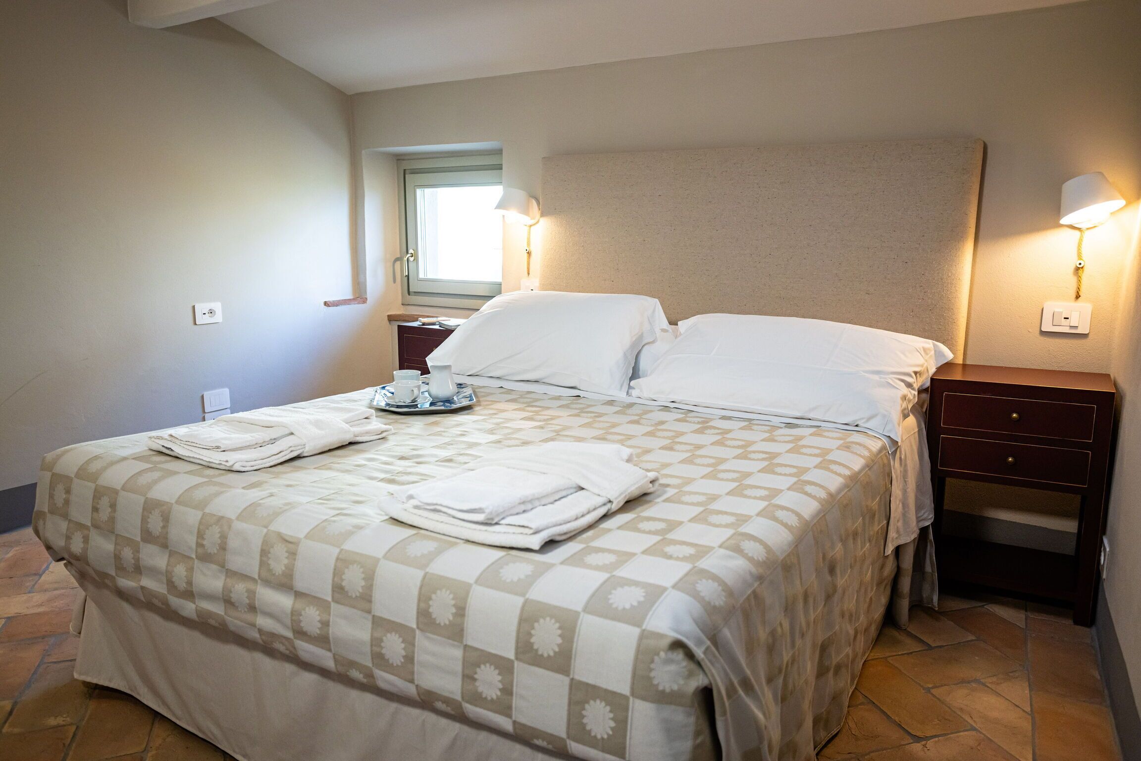 Una cama bien hecha con colcha a cuadros, dos lámparas de noche, una mesita de noche con lámpara y una ventana con luz diurna.