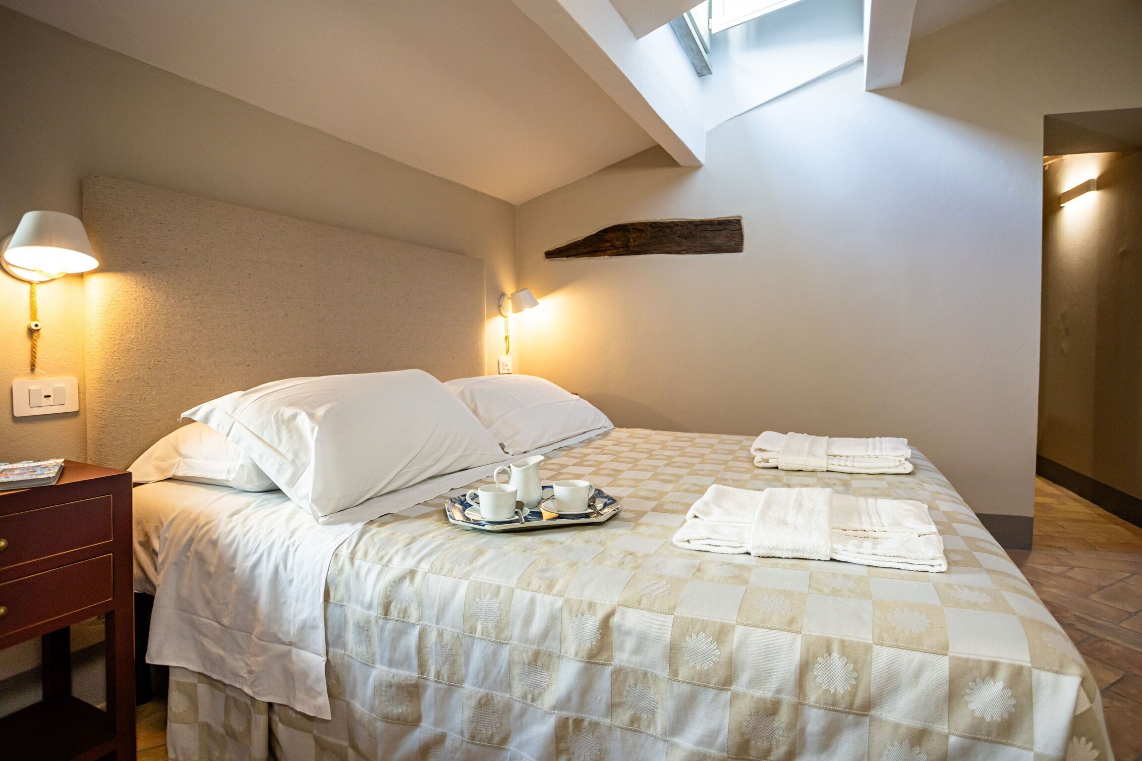 A neatly made bed with a checkered bedspread, a tray with a pitcher and cups, and towels on the bed. There is a nightstand with a lamp and a wooden headboard.