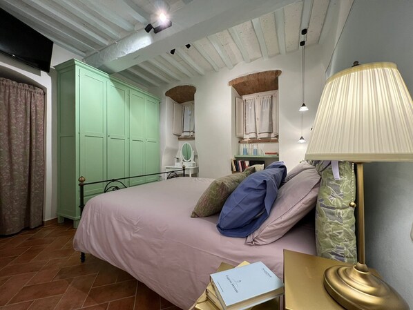 Une chambre à coucher avec un lit, une armoire verte, une coiffeuse avec un miroir, des livres et une lampe.