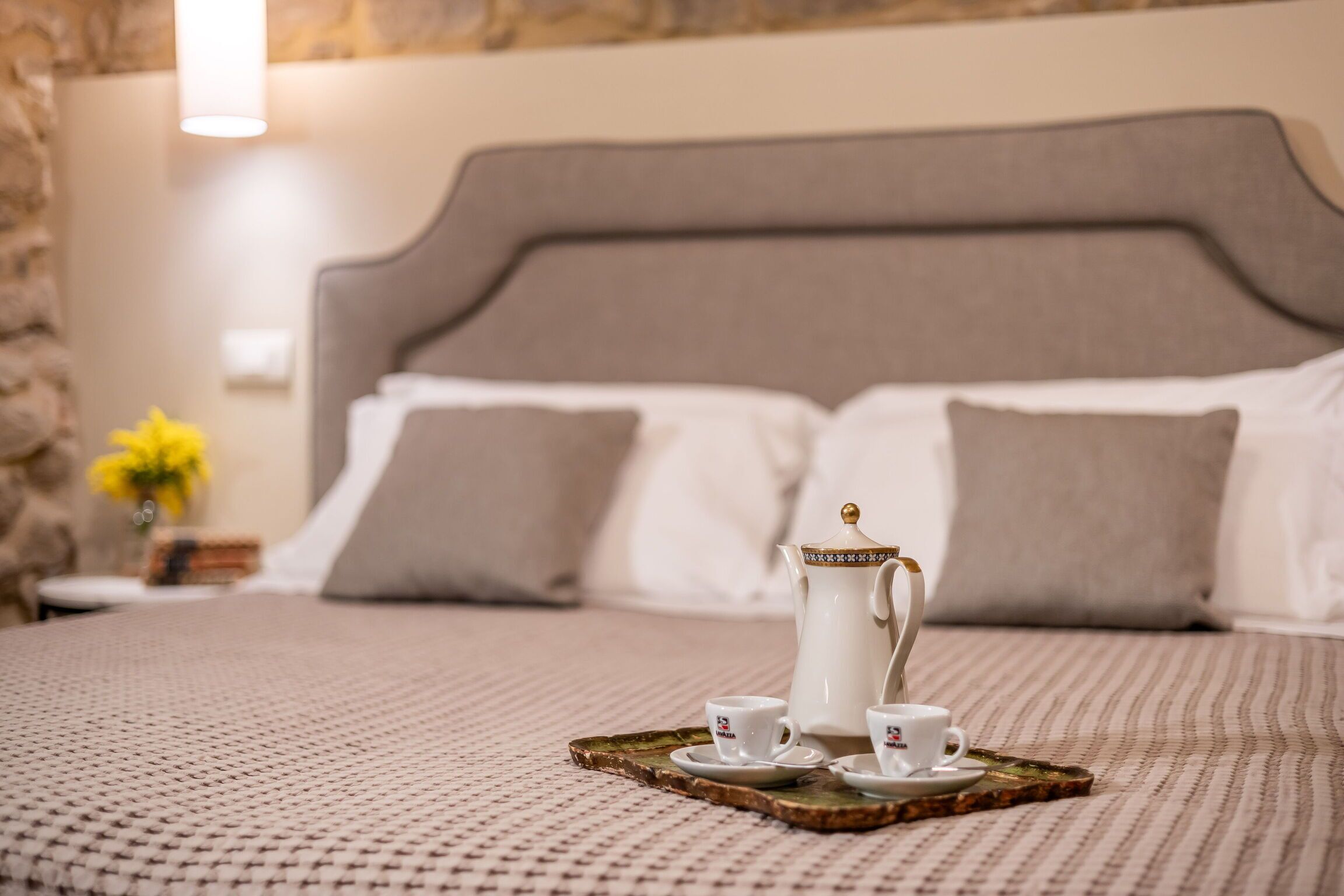 A neatly made bed with a headboard, a tray with two cups and a teapot, and a vase with yellow flowers.