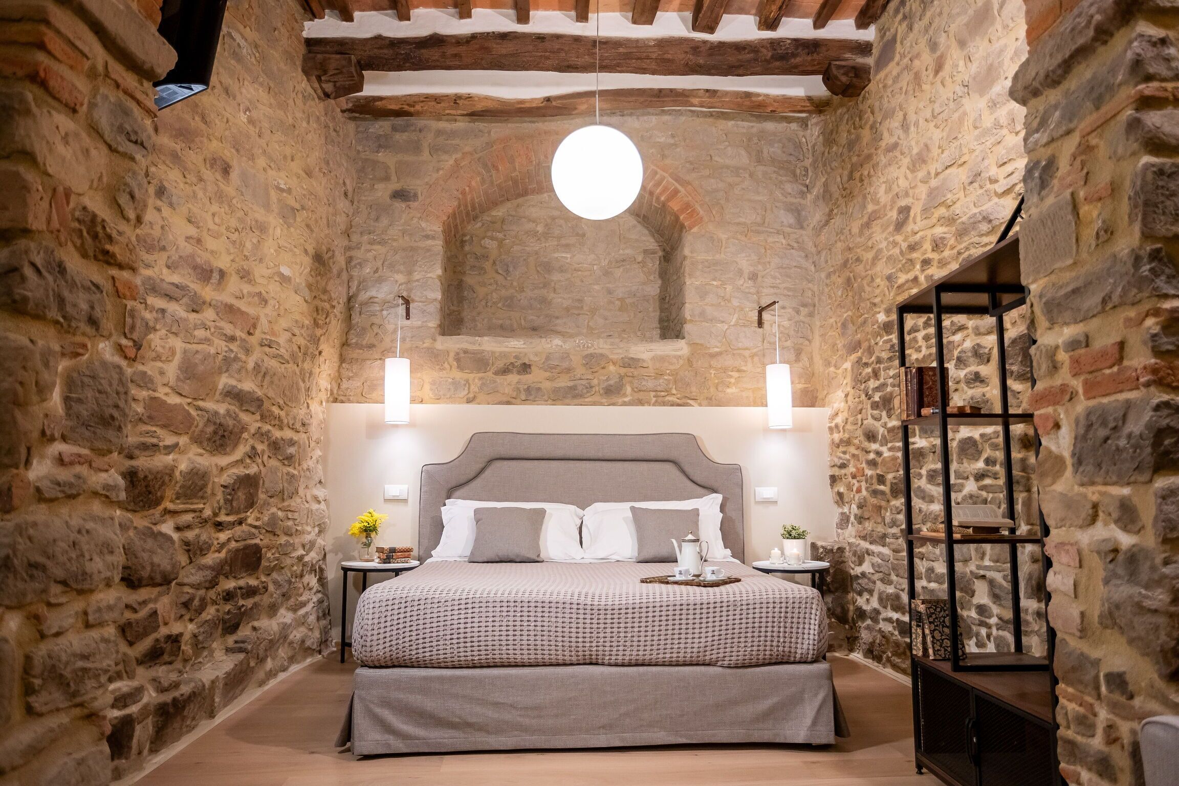 Ein Schlafzimmer mit Steinwänden, einem Bett, Nachttischen, einer Pendelleuchte und einem Holzregal.