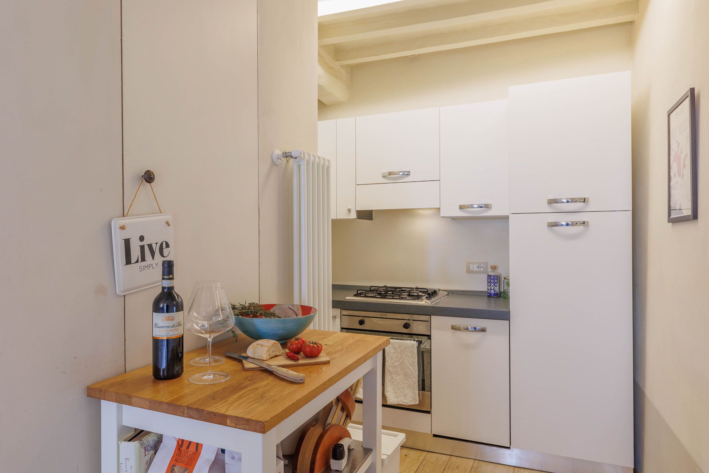 Une cuisine avec des armoires blanches, un plan de travail en bois, une bouteille de vin, un saladier de tomates et un panneau sur lequel on peut lire : « Vivez simplement ».