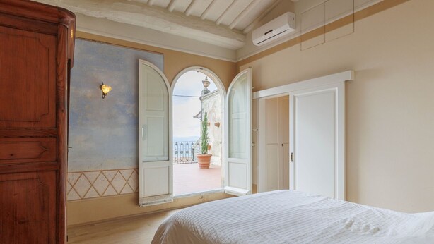 A bedroom with a large bed, a wardrobe, a mirror, and an open door leading to a balcony with a view of a building. - La Terrazza Sulla Valle (Cortona)