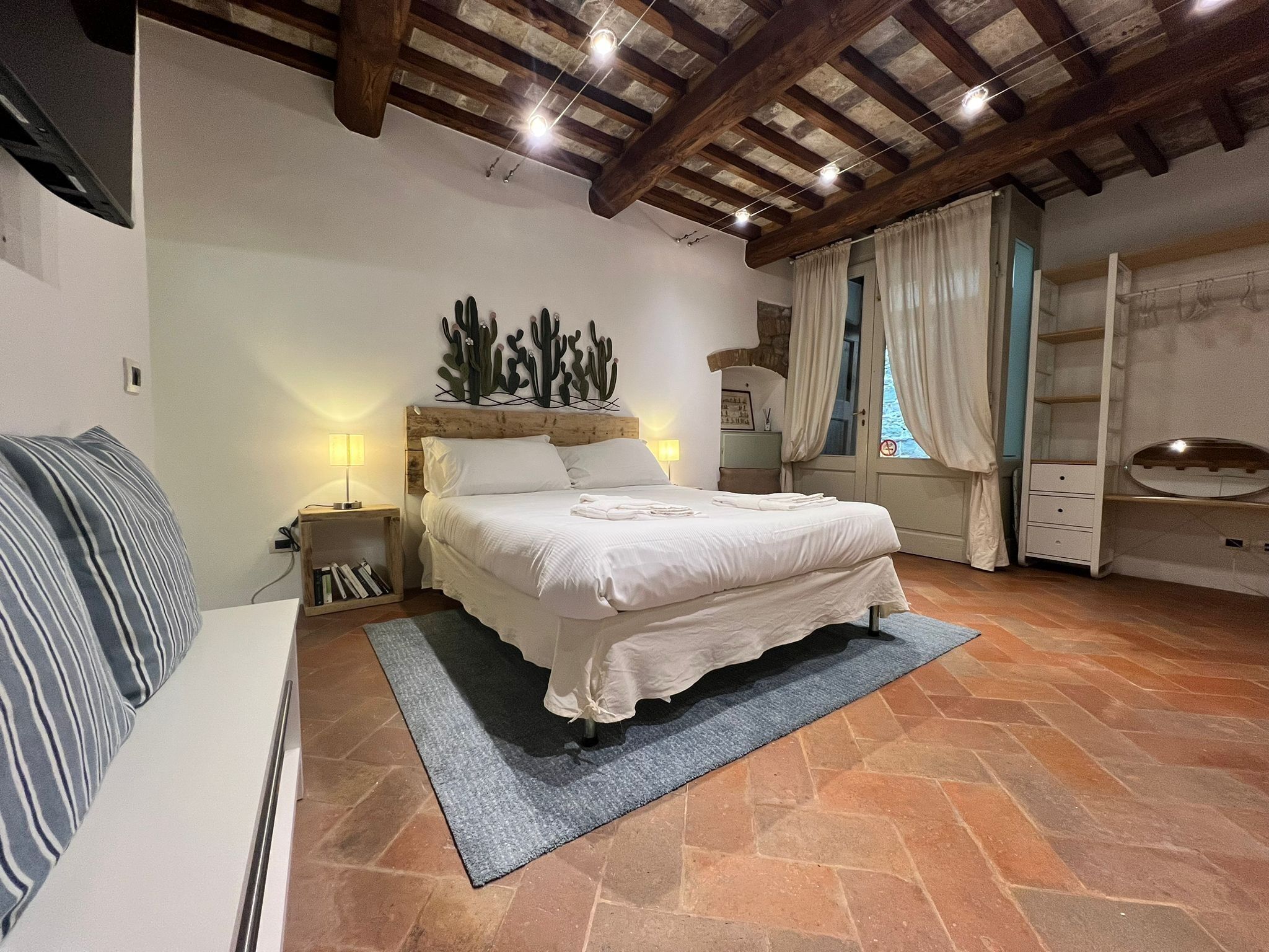 Un dormitorio con una cama grande, vigas de techo de madera, un adorno de pared con cactus, una mesita de noche con lámpara y una ventana con cortinas.