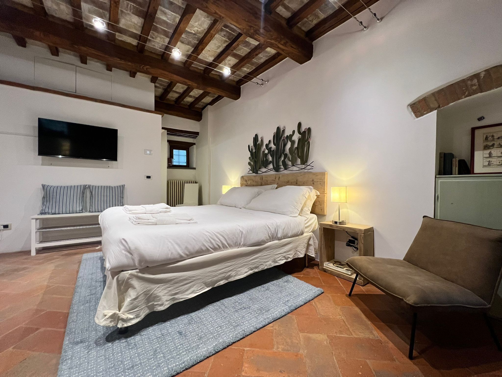 A bedroom with a bed, a nightstand, a chair, a TV, and a cactus wall art piece.