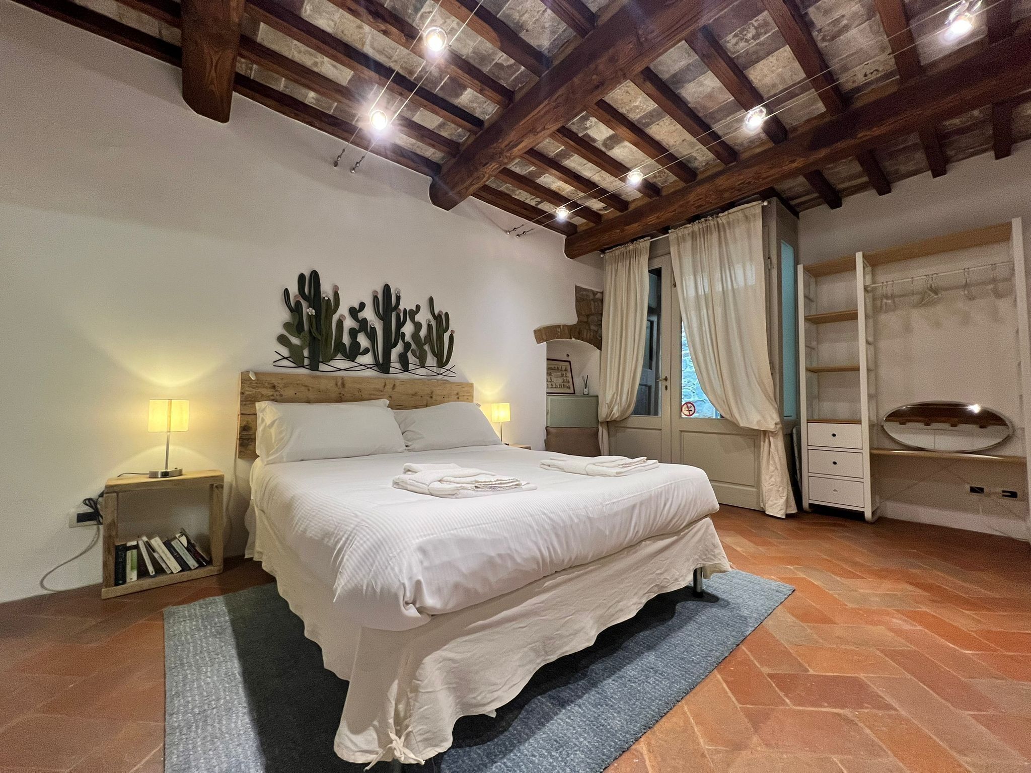 A bedroom with a large bed, wooden beams on the ceiling, a cactus wall art, a bedside table with books, and a wardrobe with a mirror.