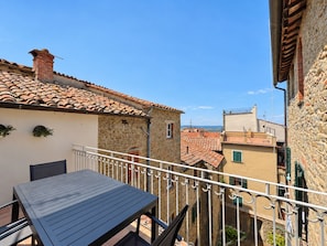 En balkon med bord og stole, der ud til en gade med traditionelle bygninger og en klar himmel.