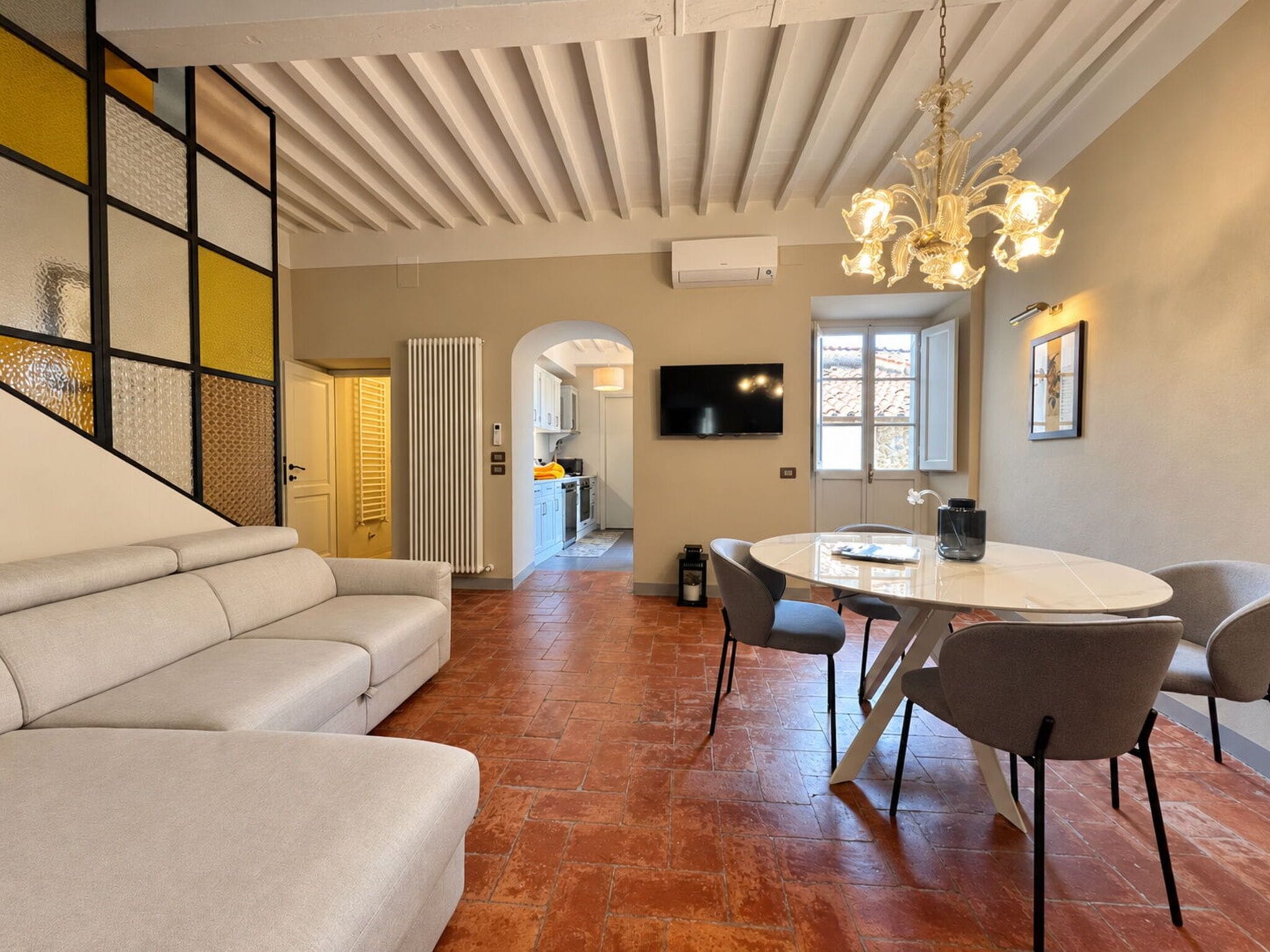 A modern living room with a sofa, a dining table with chairs, a TV mounted on the wall, and a chandelier.