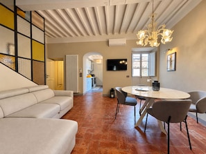 A modern living room with a sofa, a dining table with chairs, a TV mounted on the wall, and a chandelier. - Via Roma 39 (Cortona)