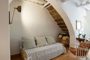 Ein gemütliches Zimmer mit einem hölzernen Bett, einem weißen Stuhl, einem Schreibtisch mit Spiegel und einem steinernen Bogen.
