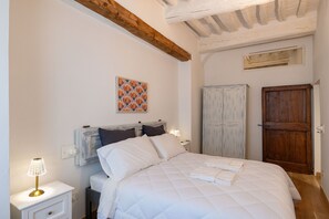 Ein Schlafzimmer mit einem weißen Bett, einem Nachttisch mit Lampe, einer Holzbalkendecke, einem Kleiderschrank und einer Tür.