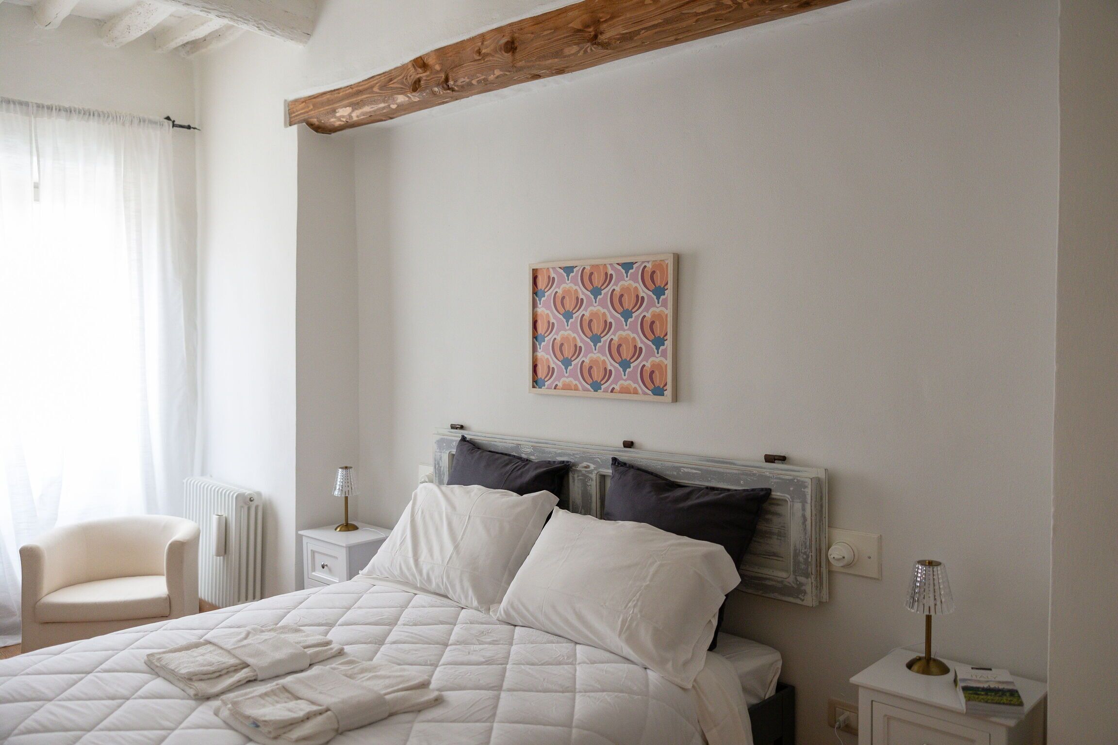 A neatly made bed with white linens, a bedside table with a lamp, a chair, and a radiator.