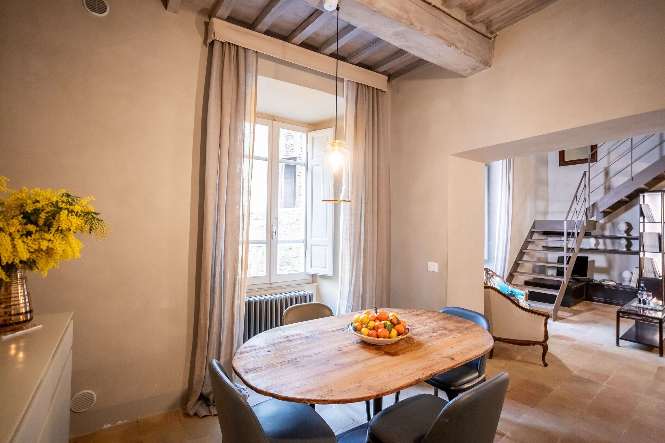 A dining area with a wooden table, chairs, a bowl of fruit, and a vase of yellow flowers.