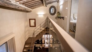 Μια σκάλα με γυάλινο κιγκλίδωμα που οδηγεί σε ένα χαμηλότερο επίπεδο. Στο ταβάνι υπάρχουν ξύλινα δοκάρια που είναι ορατά, στον τοίχο είναι τοποθετημένος ένας καθρέφτης και υπάρχει ένα ράφι με διακοσμητικά αντικείμενα.