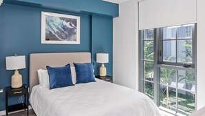 Een slaapkamer met een groot bed, twee nachtkastjes met lampen, een lijstwerk aan de wand boven het bed, en een groot raam met uitzicht op groen.