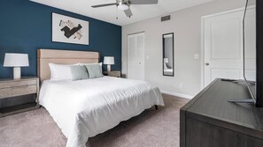 Quarto com cama, mesas de cabeceira, escrivaninha, ventoinha de teto, espelho e porta.