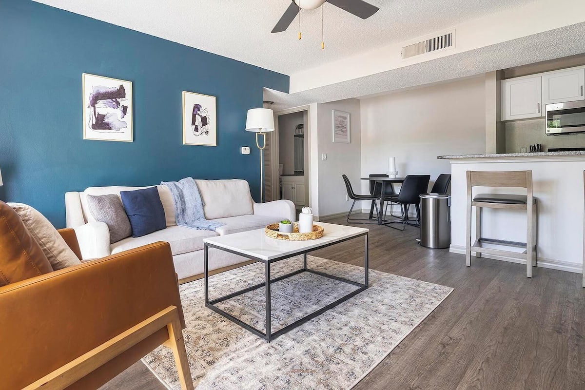 A modern living room with a blue accent wall, a leather sofa, a coffee table, and a kitchen area with a dining table and chairs.