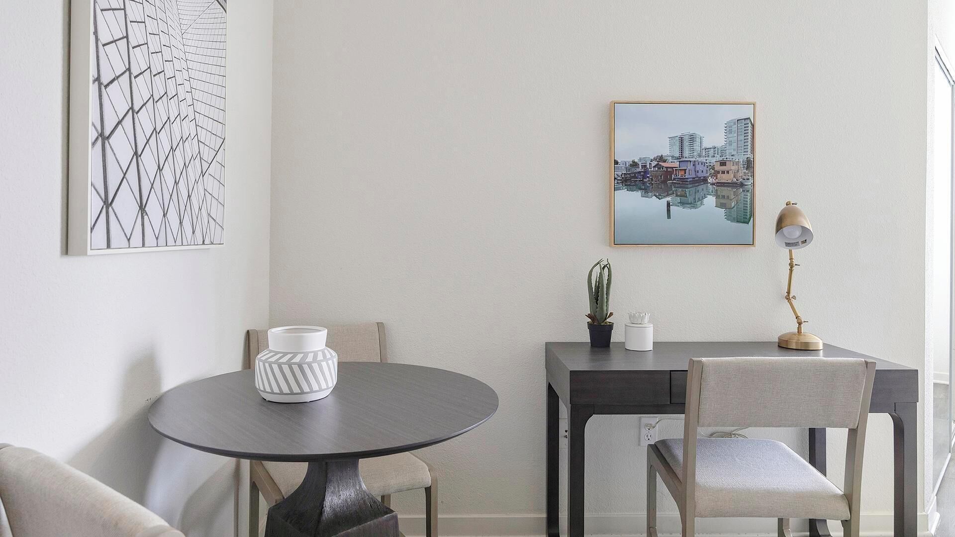 A dining area with a round table, a chair, a vase, a lamp, and a framed cityscape picture on the wall.