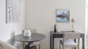 Un comedor con una mesa redonda, una silla, un florero, una lámpara y un cuadro con una vista urbana enmarcada en la pared.