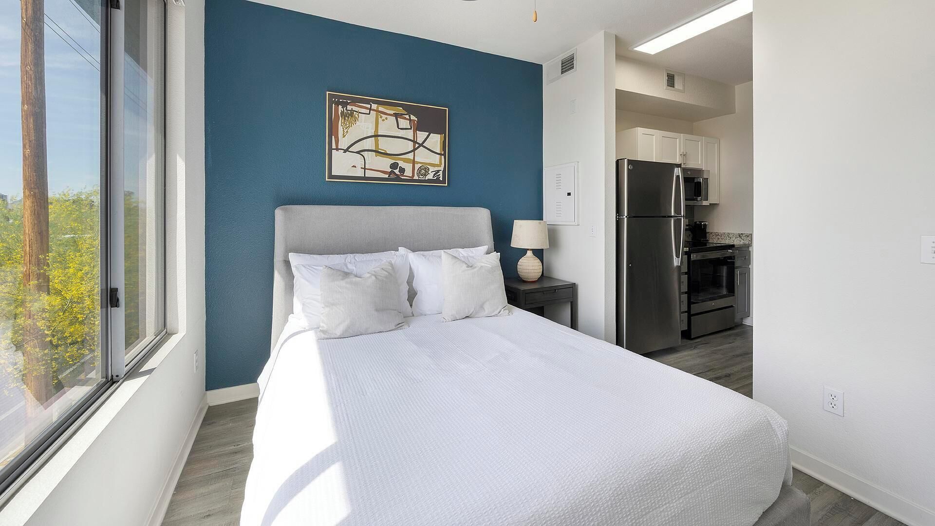Een slaapkamer met een bed, nachtkastje, lamp en een keukenhoek met een koelkast en kastjes.