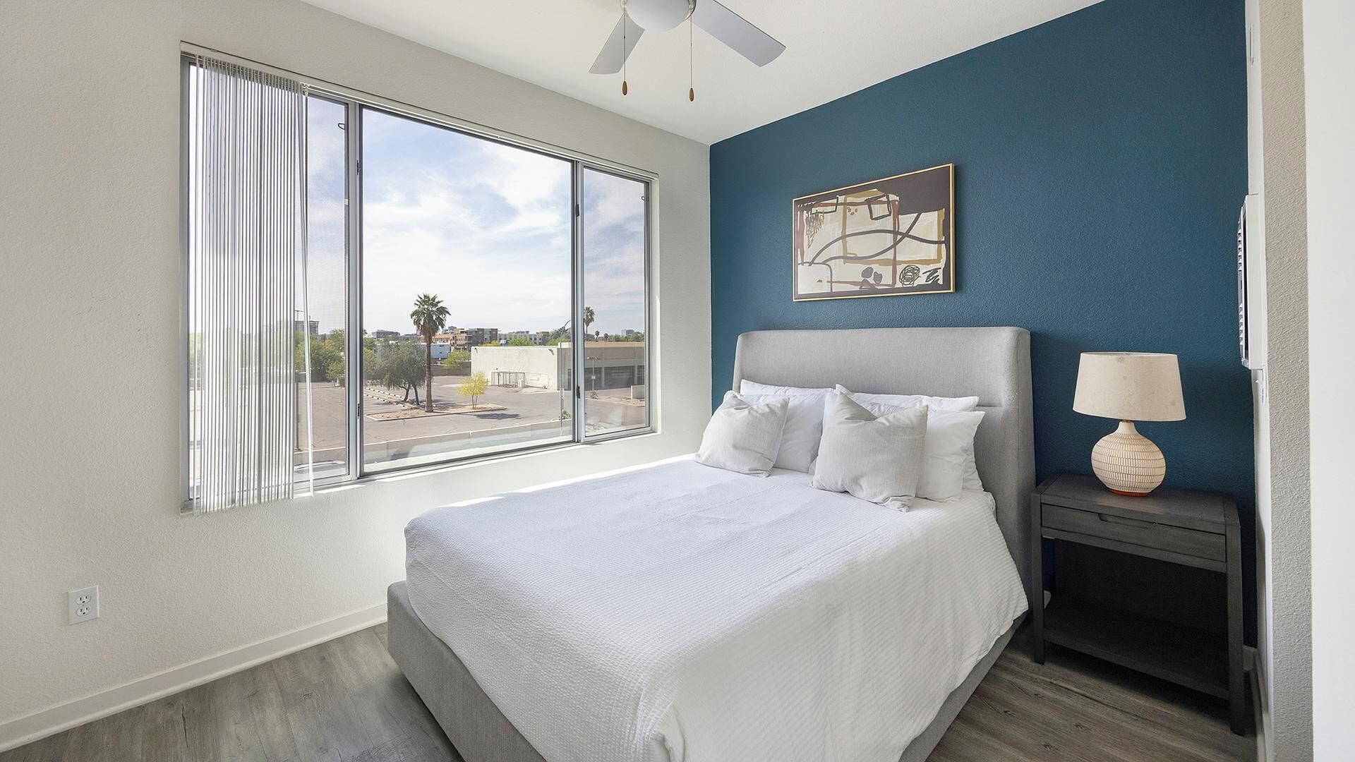 A bedroom with a large window, a bed with white bedding, a nightstand with a lamp, and a wall-mounted artwork.