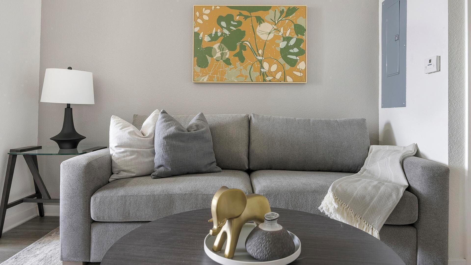 A modern living room with a grey sofa, a round coffee table with decorative items, a lamp, and a wall-mounted abstract painting.