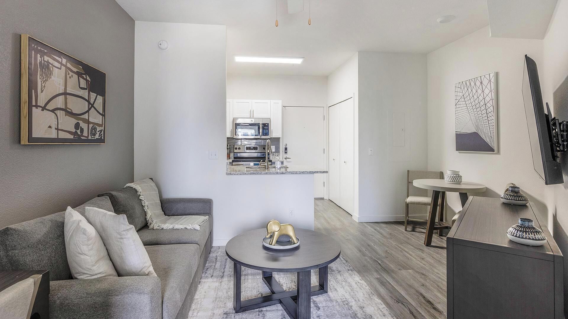 A modern apartment with a grey sofa, a round coffee table, a kitchen area with a microwave, and a dining table with chairs.