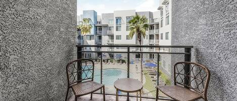 A balcony with two wicker chairs and a small round table, overlooking a swimming pool and adjacent buildings.