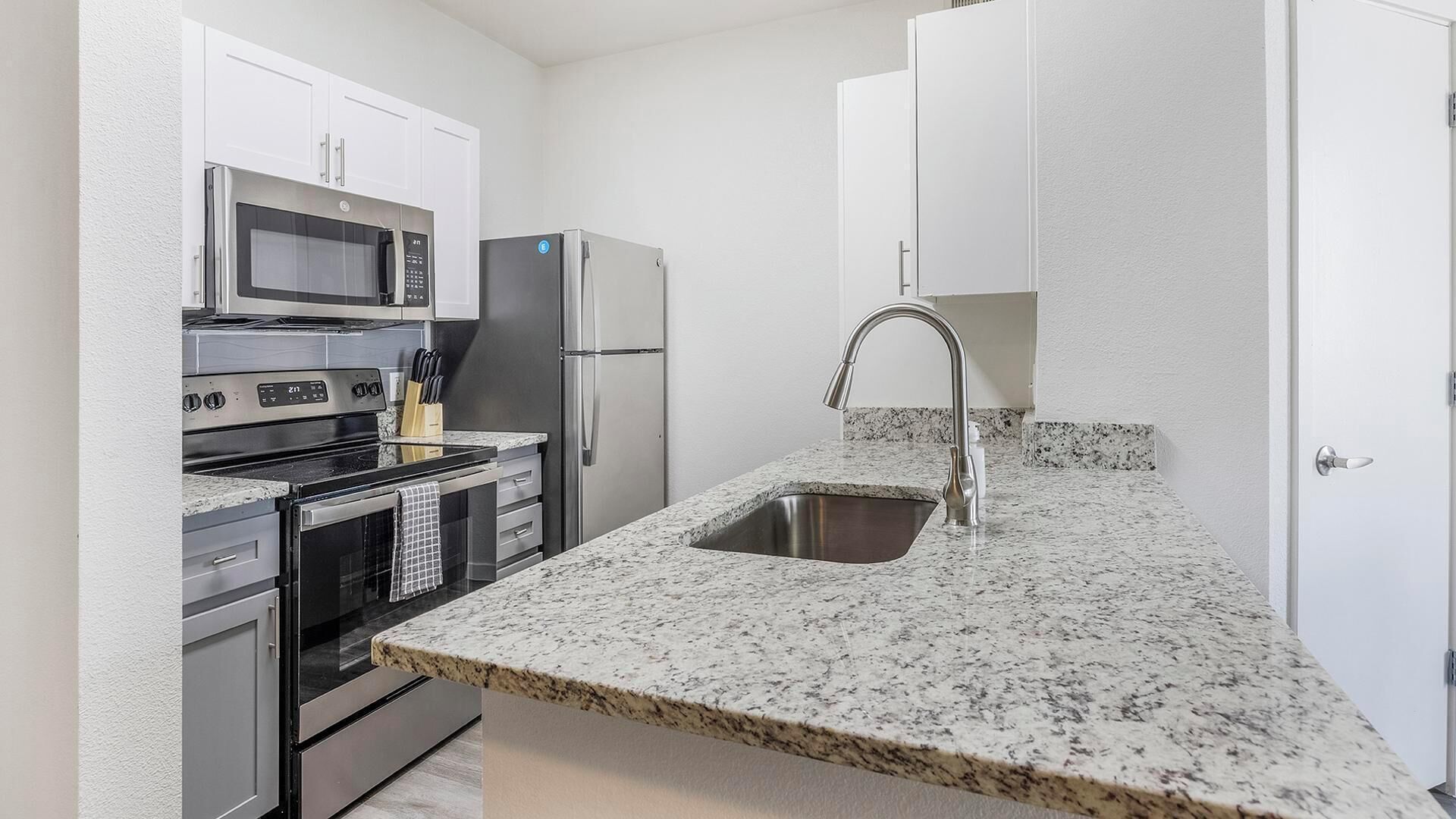 A compact kitchen with white cabinets, a stainless steel refrigerator, a microwave, an oven, and a granite countertop with a sink.