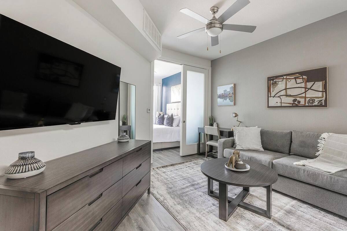 A modern living room with a flat-screen TV, a grey sofa, a coffee table, and a ceiling fan.