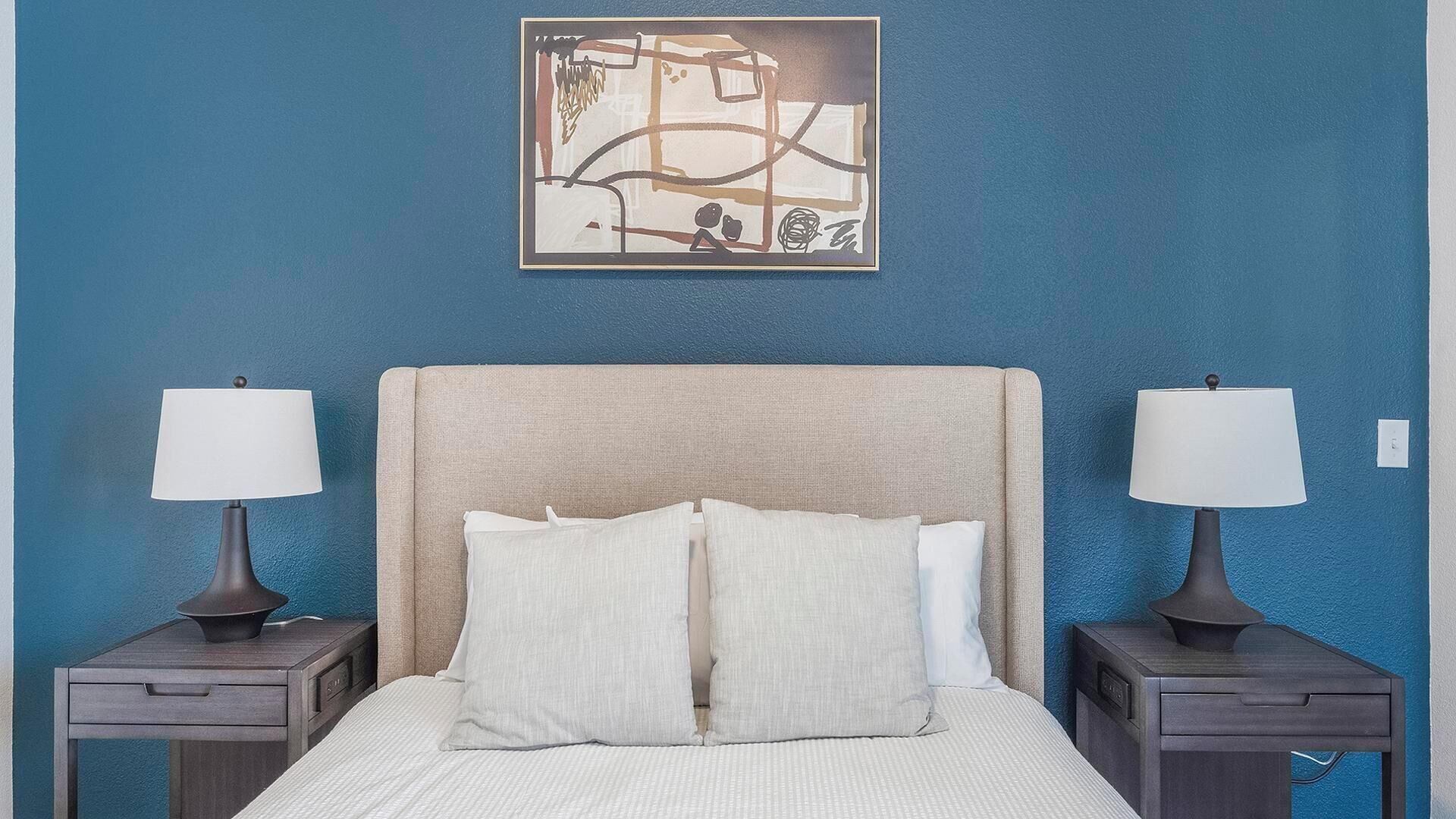 A bedroom with a large bed, two bedside tables with lamps, and a wall-mounted abstract artwork.