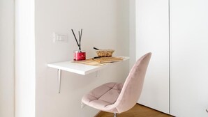 Quarto minimalista com escrivaninha branca, cadeira rosa, castiçal vermelho e cesta de fibra.