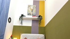 A neatly made bed with a pillow, a bedside lamp, and a wall-mounted shelf with a plant and a painting. - Lavish Partition Room in Tecom (Dubai)