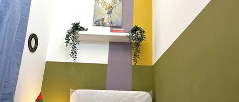 A neatly made bed with a pillow, a bedside lamp, and a wall-mounted shelf with a plant and a painting.