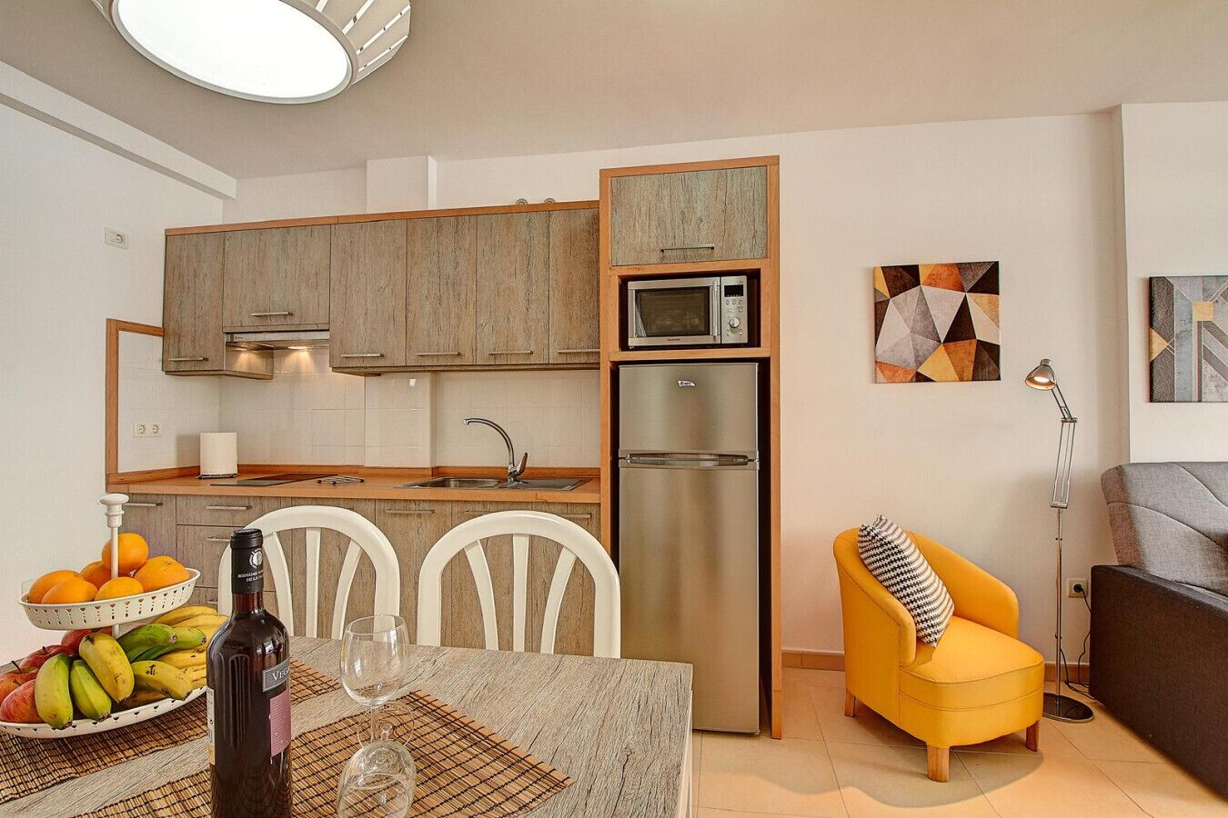 A compact kitchen with a microwave, refrigerator, and wooden cabinets. There is a dining table with chairs and a fruit basket. A yellow armchair and a floor lamp are also visible.