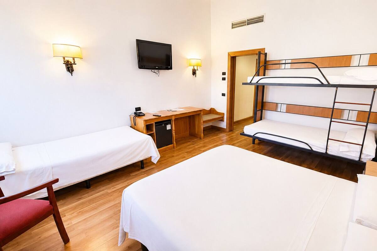 A hotel room with two single beds, a bunk bed, a desk with a chair, a television, and a lamp.